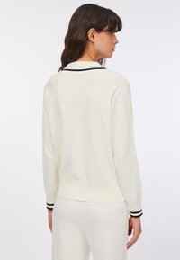Donna che indossa un maglione bianco lavorato a maglia con un motivo testurizzato sottile, colletto e polsini a righe nere, vista di spalle su uno sfondo semplice.