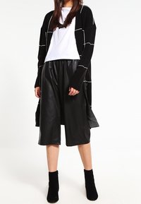 Svart lång kappa med vit rutmönster, vit t-shirt, svarta fusk-läder culotter och svarta ankelboots. Släta texturer och lagerdesign.