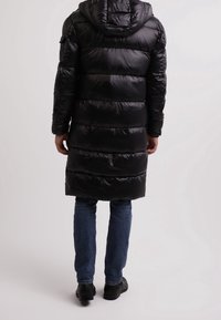 Černý prošívaný kabát s kapucí, s prošívaným vzorem. Kabát je dlouhý a lesklý, doplněný modrými džínami a černou obuví.