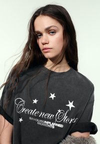 Oversized t-shirt i kulgrå med hvide stjerneaccenter og tekst i skrift, der lyder "Create new Stories" og "Savour unplanned Moments."