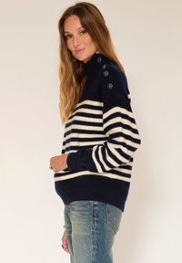 Pull rayé bleu marine et crème avec un col montant et un détail de quatre boutons sur l'épaule. Matériau doux et coupe décontractée.