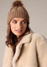 Bruine gebreide muts met een textuurpatroon en een fluffy pompon bovenop, gecombineerd met een beige kunstbontjas. Close-up van de muts en schouders.