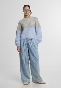 Fiatal nő világoskék és szürke kötött pulóverben, széles szárú világoskék farmernadrágban, bézs sportcipőben és áttetsző szemüvegben áll a fehér háttér előtt.