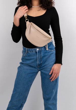 Bezová kožená crossbody taška s zakřiveným tvarem, zlatými kovovými doplňky a zipovým uzavřením. Stylizována s černým topem a modrými džínami s vysokým pasem.