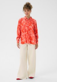 Frau mit lockigem Haar, die ein orangefarbenes Hemd mit Blumenmustern, cremefarbene, weite Hosen und rote Schuhe trägt, steht vor einem schlichten Hintergrund.