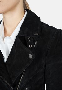 Veste de motard en suède noir avec un col cranté, des éléments en argent, une fermeture éclair et des accents à boutons, présentant une finition douce et texturée.