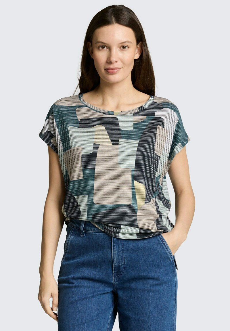 Femeie cu păr lung și închis la culoare, purtând un top cu mâneci scurte, cu un model abstract multicolor, și blugi albaștri, având o mână în buzunar.