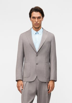 Homme portant une veste de costume gris clair et un pantalon assorti par-dessus une chemise à boutons bleu pâle sur un fond blanc uni.