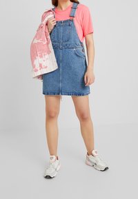 Denim overall dress with adjustable straps, blue color, front pockets, and a front button closure, paired with a pink tie-dye jacket and white sneakers.
