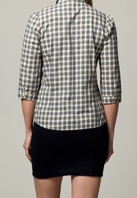 Donna che indossa una camicia a scacchi blu, bianca e beige con maniche a tre quarti e una gonna nera aderente, vista di spalle.