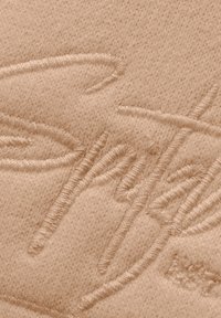 Beige stof met verhoogde borduurwerk van een gestileerd logo, met een zachte textuur en een glad oppervlak met minimale patroondetails.