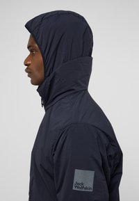 Marineblaues Kapuzensweatshirt mit hohem Kragen, leichtem Material, flachen Nähten und einem Logopatch am Ärmel. Seitenansicht.