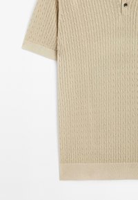 Beige kortærmet polo med et struktureret kabelstrik mønster, med en placket med tre knapper og ribkant ved kant og ærmer.