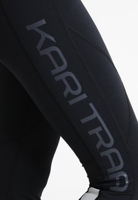 Černé legíny s hladkou texturou, s výrazným šedým logem "KARI TRAA" po straně a kontrastními bílými detaily na lemu.