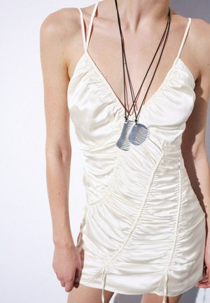 Mujer que lleva un vestido lencero de raso blanco fruncido con tirantes finos y dos colgantes grises con forma de peine en cordones negros alrededor del cuello.