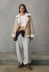 Giacca in shearling beige con colletto bianco, top corto bianco, pantaloni sportivi grigi e scarpe da ginnastica marroni con lacci decorati. Abbigliamento streetwear semplice e rilassato.
