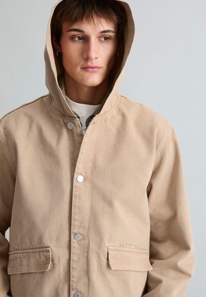 Junge Person trägt beige Kapuzenjacke mit Druckknöpfen, schaut seitlich vor einem schlichten hellen Hintergrund.