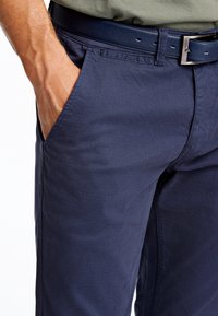 Męska dłoń spoczywa w przedniej kieszeni granatowych spodni o fakturze, z pasującym skórzanym paskiem i włożoną do środka oliwkowozieloną koszulą.