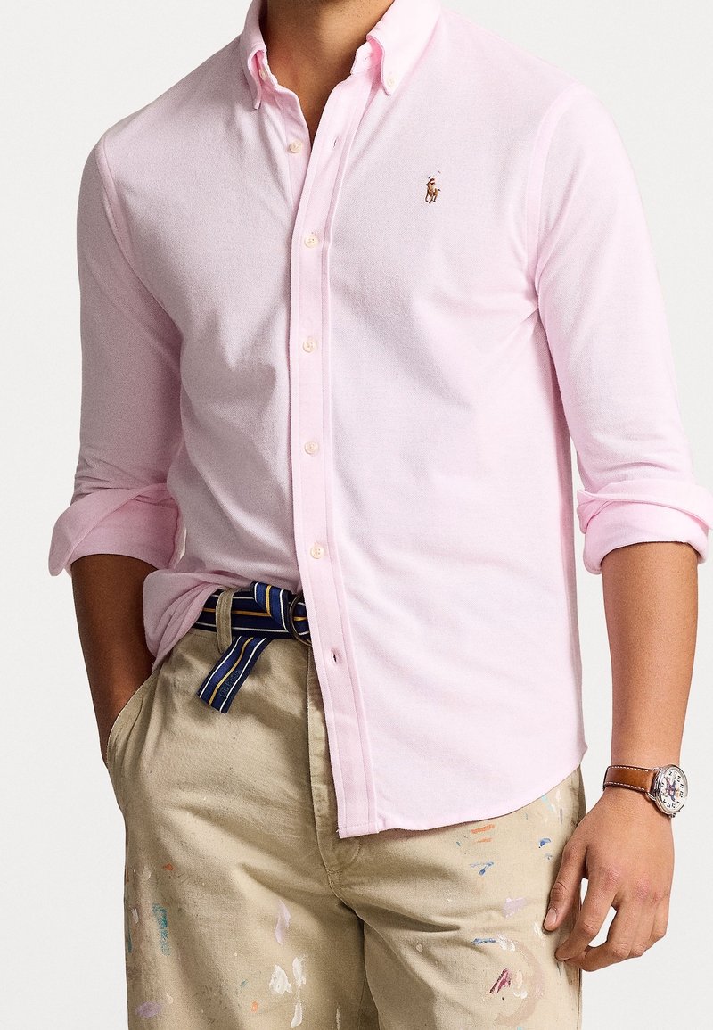Ljusrosa skjorta med knappar och uppvikt ärm, prydd med en liten broderad logotyp. Bärs med beige byxor och ett randigt bälte.