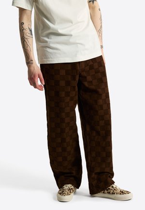 Person iført brune ternede vidde ben bukser, hvid t-shirt og hvide sneakers med sorte pletter, stående mod en ensfarvet baggrund.