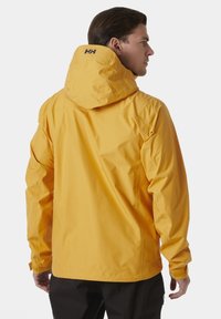 Geel waterproof jack met een capuchon, voorzien van een matte afwerking, elastische manchetten en logo-detail op de achterkant. Ontworpen voor gebruik buitenshuis.