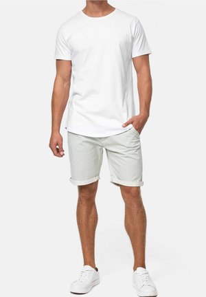 Mand iført en simpel hvid kortærmet t-shirt, lysegrå ternede shorts med opkrogede ben og hvide sneakers, stående med den ene hånd i lommen.