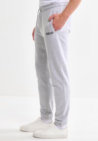 Uomo che indossa pantaloni della tuta grigio chiaro con polsini elasticizzati, calzini bianchi e scarpe da ginnastica bianche, in piedi con una mano in tasca.