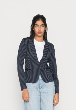 Blazer blu navy a righe con un design aderente, chiusura a un bottone e rever a punta, abbinato a una t-shirt bianca e jeans blu chiaro.
