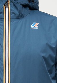 Μπλε μπουφάν K-Way με φερμουάρ ρίγας σε λευκό, ναυτικό μπλε και κίτρινο, με το λογότυπο K-Way στο στήθος και κουκούλα προσαρτημένη.