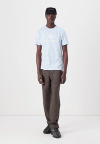 Lichtblauw katoenen T-shirt met "BELSTAFF" logo, gecombineerd met losse bruine broeken en zwarte sneakers. Een zwarte pet maakt de outfit af.