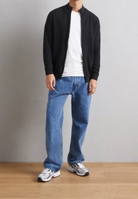 Zwarte ritscardigan over een wit T-shirt, gecombineerd met blauwe losse jeans en grijze sportschoenen op een houten vloer.