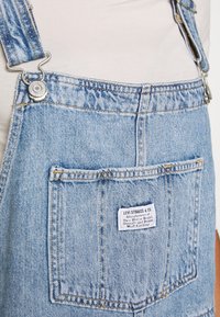 Ljusblå denimoverall med justerbara metallclips, frampocket och en vävd etikett med tillverkningsuppgifter. Texturerad tyg.