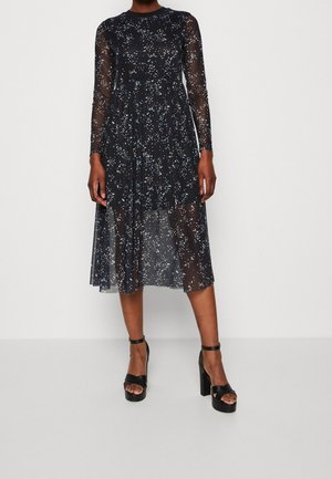 Kvinde iført en sort gennemsigtig midi-kjole med lange ærmer og lyseblå og hvide prikker, kombineret med sorte højhælede platformsandaler.