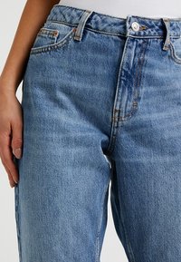 Los jeans de mezclilla azul claro presentan una cintura alta, corte recto y costuras naranjas en contraste. Tienen un estilo clásico de cinco bolsillos.
