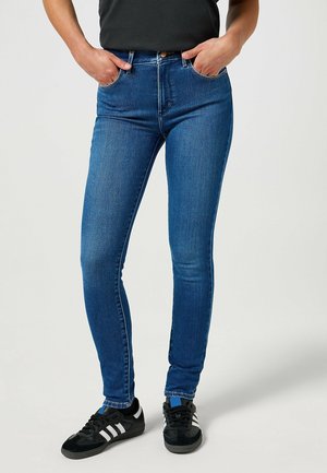 Benen in nauwsluitende blauwe spijkerbroeken en zwarte sneakers met witte strepen, handen in de voorzakken van de spijkerbroek gestoken tegen een effen achtergrond.