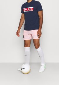 Modré tričko s červeným logem "PARIS", růžové šortky, bílé ponožky Nike a bílý fotbalový míč s černými detaily na šedém pozadí.