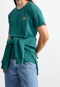 Turkusowy bawełniany t-shirt z okrągłym dekoltem i krótkimi rękawami, z małym pomarańczowym logo. Sweter związany w talii na dżinsach.