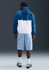 Homme aux cheveux tressés portant une veste à capuche bleue et blanche, un short cargo en denim clair, des chaussettes blanches et des baskets blanches, debout devant un fond sombre.