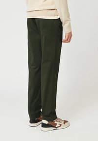 Persona che indossa pantaloni verde scuro, un maglione beige e scarpe da ginnastica marroni e beige, in piedi contro uno sfondo neutro.
