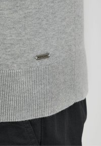 Maglione grigio lavorato a maglia con orlo a costine, caratterizzato da una piccola etichetta metallica. Tessuto strutturato, aspetto morbido, abbinato a pantaloni scuri.