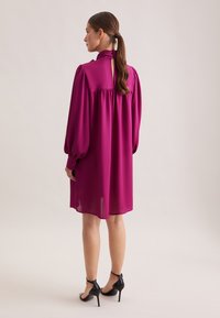 Abito fucsia con maniche lunghe a sbuffo e dettaglio a chiave sul retro, realizzato in un materiale leggero. Sandali neri con tacco alto completano il look.