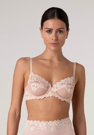 Donna con capelli scuri che indossa un reggiseno in pizzo rosa chiaro e mutandine in pizzo abbinate a vita alta, in piedi davanti a uno sfondo semplice.