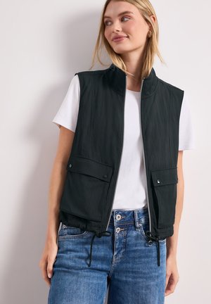 Junge Frau trägt eine schwarze Weste mit Reißverschluss und Fronttaschen über einem weißen T-Shirt und blauen Jeans, schaut seitlich mit einem leichten Lächeln.