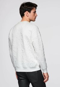 Sweat-shirt blanc texturé avec motif de lettres en relief, col rond et poignets côtelés. Porté avec un jean noir, vu de dos.