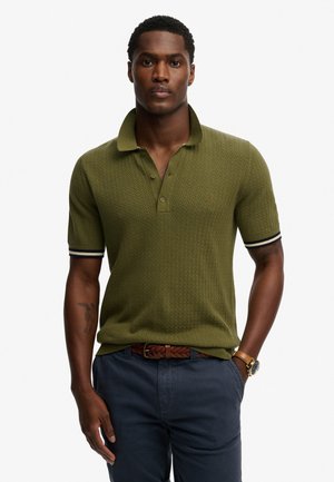 Korte mouwen polo shirt in olijfgroen met eenTextuurpatroon en een knoopsluiting met twee knoppen. Kenmerkt zich door contrasterende gestreepte manchetten, gecombineerd met marineblauwe broek.