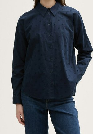 Vrouw draagt een marineblauwe blouse met knopen en getextureerde polkadots en blauwe jeans, poseert met één hand in de zak.