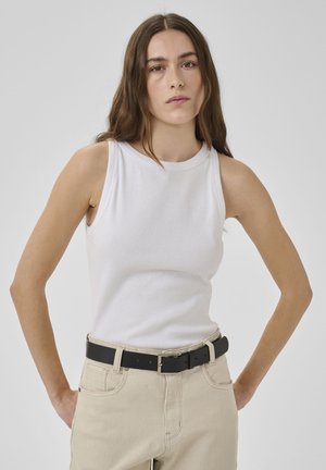 Young woman with long brown hair wearing a white sleeveless top, beige pants, and a black belt, standing against a plain gray background.
