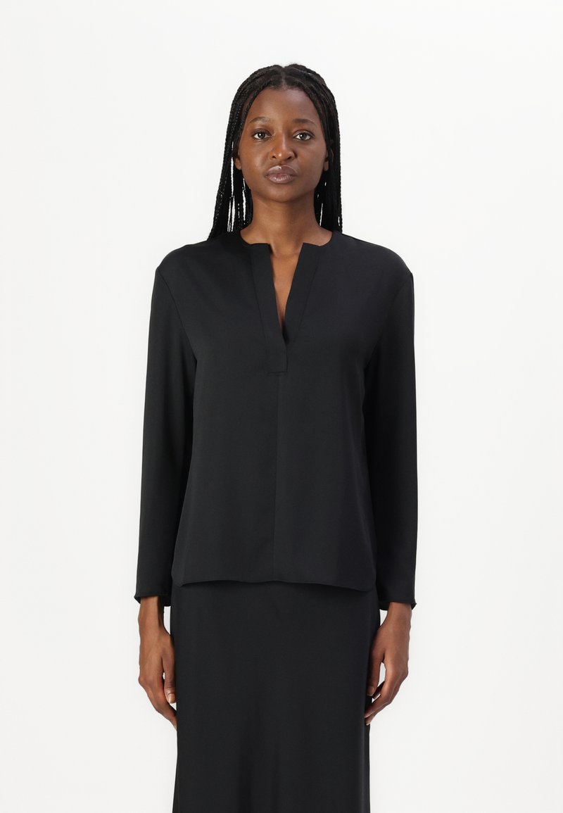 Blusa nera a maniche lunghe con scollo a V e taglio dritto, realizzata in tessuto morbido; indossata con una gonna abbinata, caratterizzata da un design minimalista.