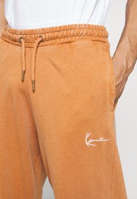 Orange cotton sweatpants with an elastic waistband, drawstring, and side pockets. Features a white embroidered logo on the left thigh.