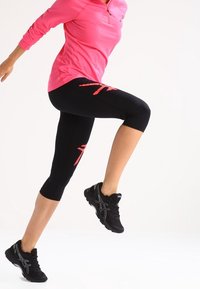 Person, die ein pinkes Langarmshirt, schwarze knielange Leggings mit roten Akzenten und schwarze Laufschuhe trägt, hebt ein Knie in einer dynamischen Pose.